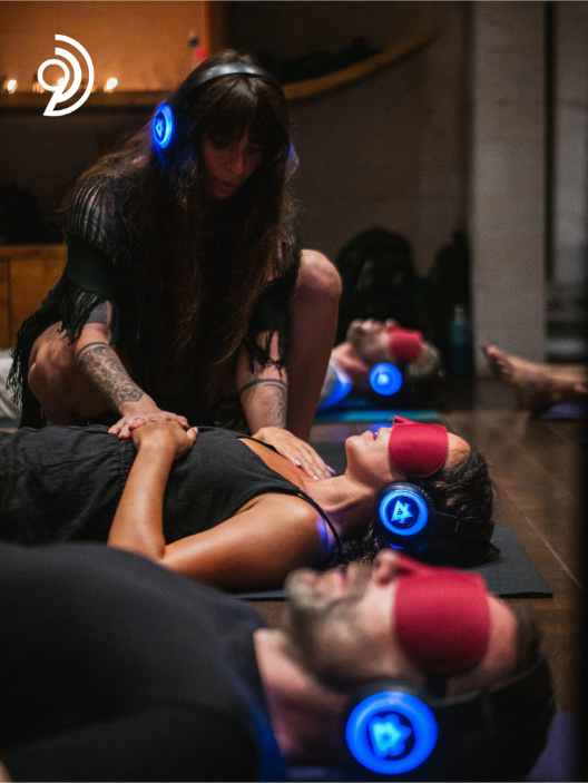 9D Breathwork Session image, from Breathwork Coach in Barrie, where someone is crouched down with hands on someones stomach and shoulder while they are laying on the floor with others with eye coverings on and a headset