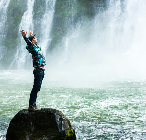 Life Coaching Services Image of man with arms raised to the sky standing on a rock in front of a waterfall